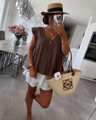 A woman wearing a brown sleeveless V-neck top, white ruffled shorts, a straw hat, and carrying a beige woven tote bag 