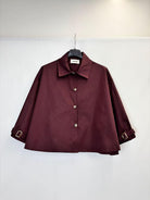 A dark burgundy long-sleeve trench jacket with a collar and decorative silver-tone buttons, hanging on a black hanger against a plain white wall.