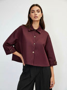 A woman modeling a cropped, long-sleeve button-up jacket in deep burgundy with a collar and buttons, paired with black trousers,