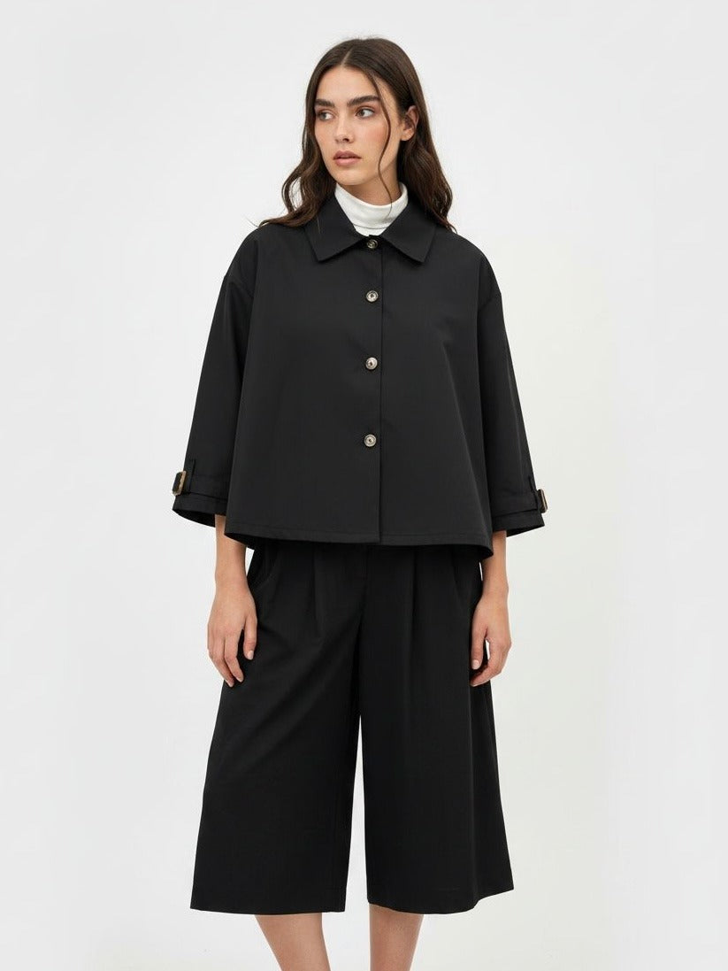 A model wearing a black cropped button-up jacket with three-quarter sleeves, paired with matching black wide-leg cropped pants and a white turtleneck underneath