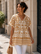 A woman wearing a beige and white patterned short-sleeve tunic top with white pants, walking outdoors on a city sidewalk next to a beige stone building.