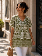 A woman wearing a short-sleeve V-neck tunic top with an olive green and white bohemian-style pattern, paired with white pants and carrying a woven straw handbag, walking outdoors on a city street.