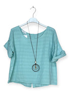 A light aqua blue short-sleeve top with a relaxed fit, hanging on a white hanger with a long black pendant necklace draped over it, against a plain white background.