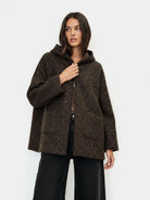 A woman modeling a dark brown textured hooded jacket with a fuzzy boucle-like pattern, paired with black wide-leg pants