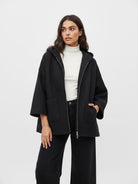 A woman modeling a black textured hooded zip-up jacket with wide sleeves, worn over a white turtleneck and black wide-leg pants