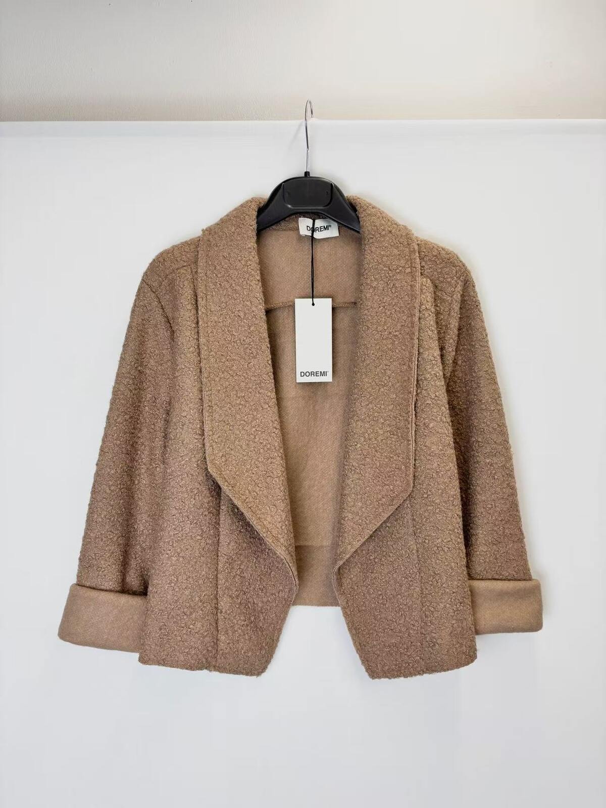 A tan textured bouclé open-front jacket with wide lapels and rolled cuffs, hanging on a black hanger 