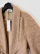 A close-up of textured bouclé coat in a warm camel beige color, hanging on a black hanger 