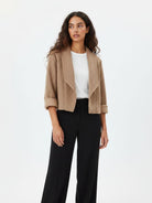 A woman modeling a cropped, open-front beige bouclé jacket with rolled cuffs over a white top and black wide-leg trousers, photographed against a clean white studio background.