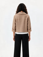 A model shown from the back wearing a cropped beige jacket with rolled three-quarter sleeves, black wide-leg trousers, and a white undershirt, against a plain white studio background.