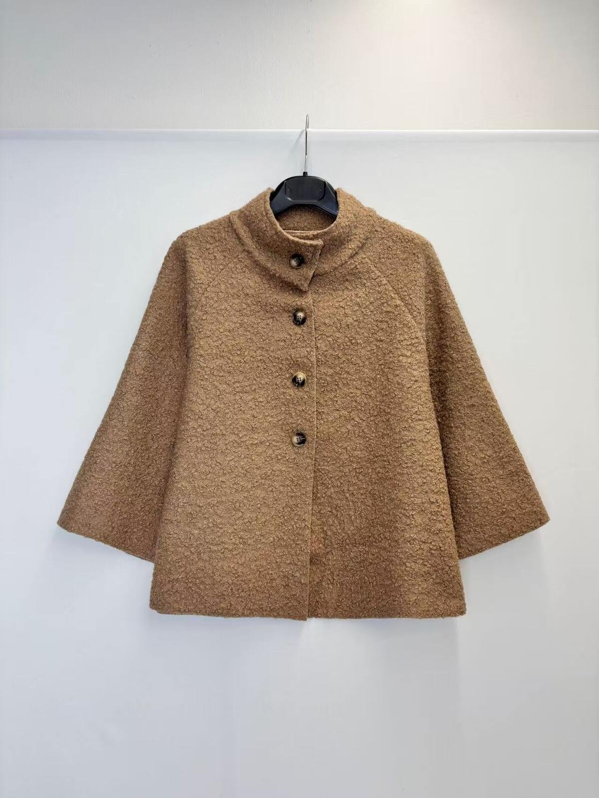 A brown textured bouclé jacket with a mock neck and three-quarter sleeves, hanging on a black hanger against a plain white wall.
