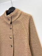 A close-up of a textured, bouclé-like brown jacket hanging on a black hanger against a plain white wall.