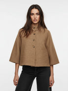 A woman modeling a cropped, high-neck, short-sleeved cape jacket in a textured camel brown fabric, paired with black jeans, against a plain white studio background.