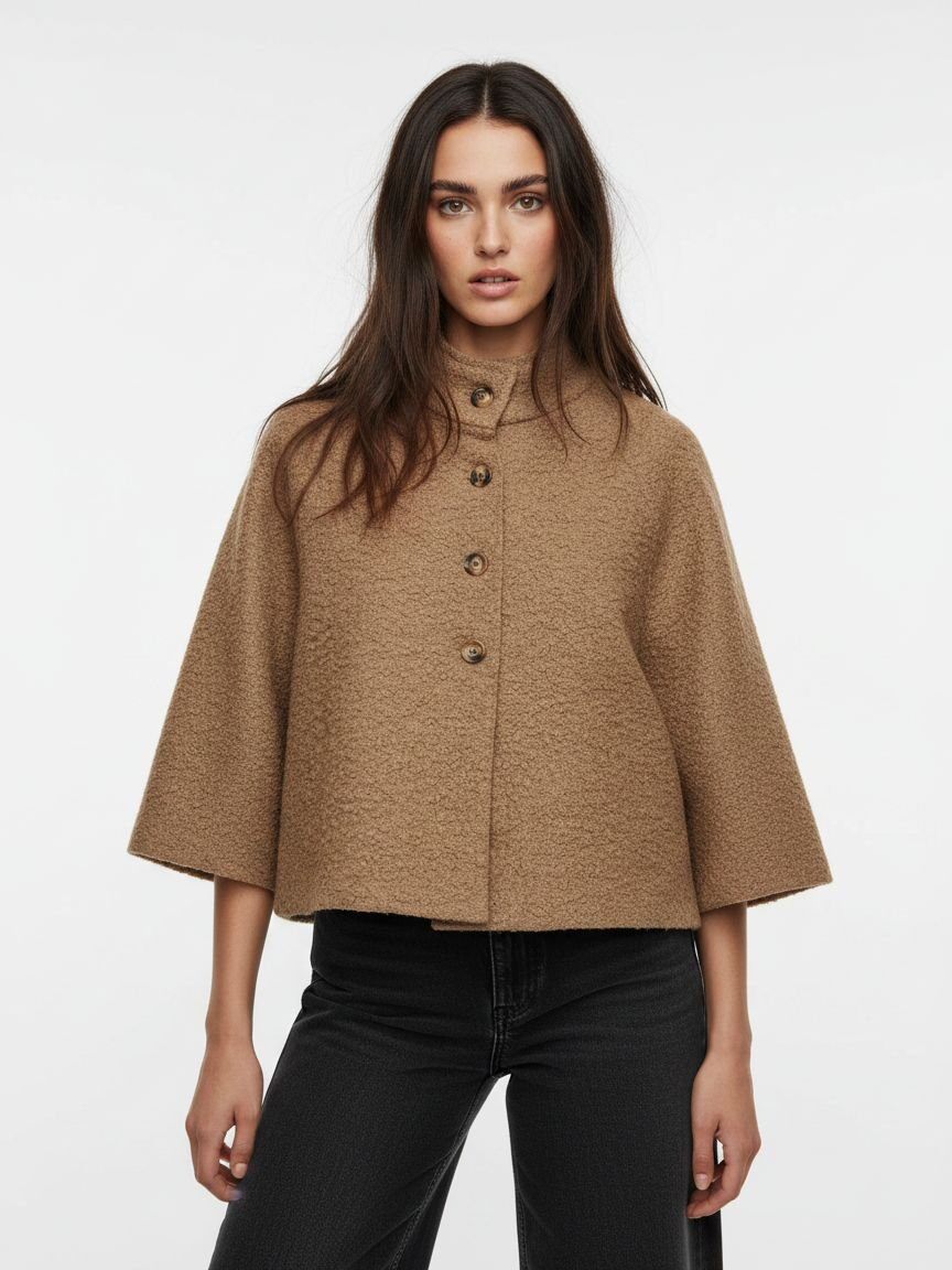 A woman modeling a cropped, high-neck, short-sleeved cape jacket in a textured camel brown fabric, paired with black jeans, against a plain white studio background.