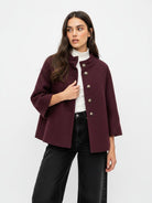A woman modeling a short, textured burgundy jacket with a high stand collar and decorative silver-tone buttons, worn over a white turtleneck and black wide-leg jeans, against a plain white studio background.