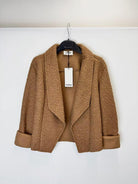 A camel brown bouclé cropped jacket with wide lapels and rolled cuffs, hanging on a black hanger 