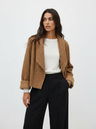 A woman modeling a cropped, open-front teddy fleece jacket in camel brown, worn over a white ribbed top and black wide-leg trousers, against a plain light gray studio background.