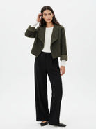 A woman modeling a cropped olive green textured jacket with rolled cuffs, worn over a white long-sleeve top and black wide-leg trousers, against a plain white studio background.