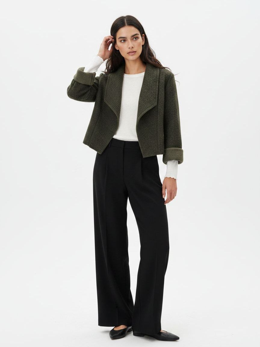 A woman modeling a cropped olive green textured jacket with rolled cuffs, worn over a white long-sleeve top and black wide-leg trousers, against a plain white studio background.