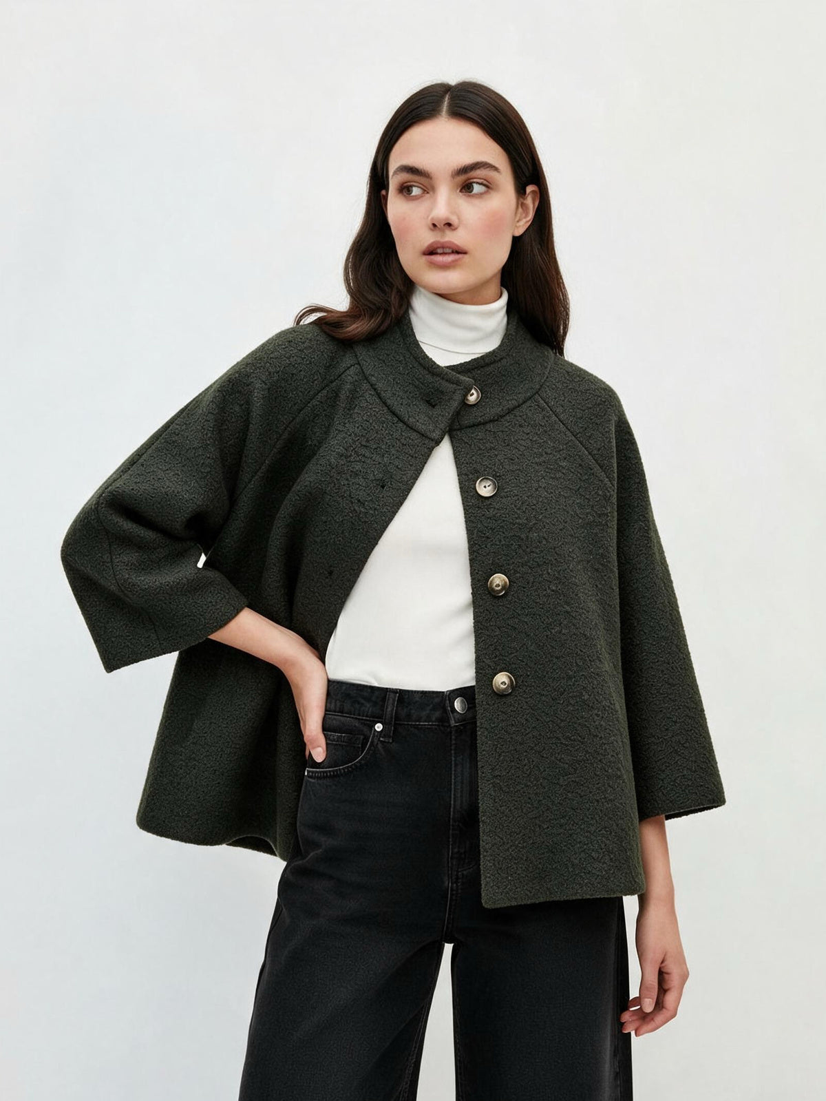 A woman modeling a dark olive green textured cropped jacket with wide three-quarter sleeves and gold-tone buttons, worn over a white turtleneck and black jeans against a light gray studio background.