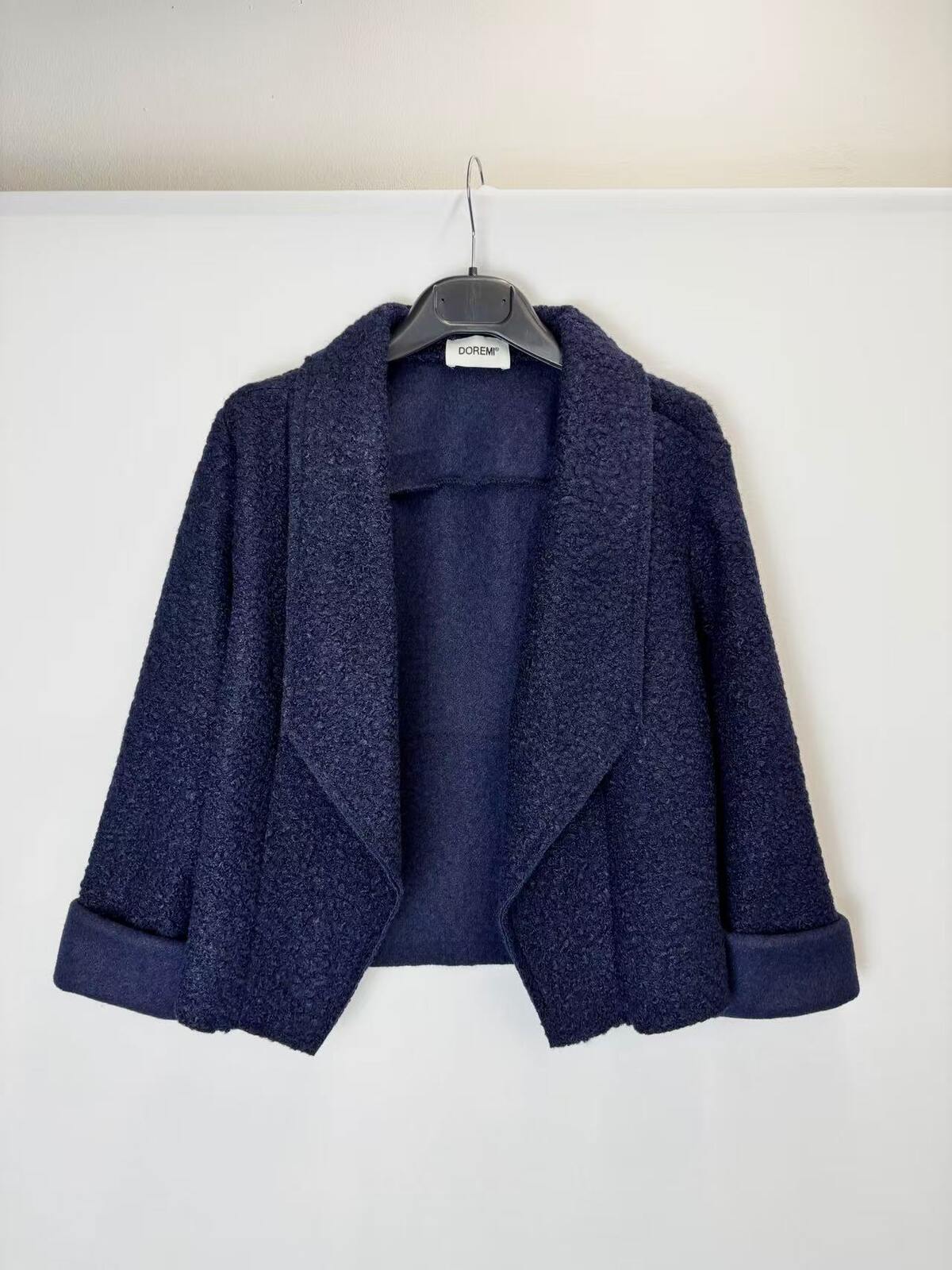 A navy blue textured bouclé jacket with wide lapels and rolled cuffs, hanging on a black hanger against a plain white wall.