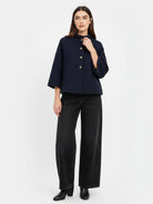 A model wearing a dark navy blue cropped jacket with three-quarter sleeves and gold buttons, paired with black wide-leg trousers and black boots, against a plain white studio background.