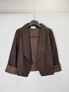 A brown textured open-front jacket with wide lapels and rolled cuffs, hanging on a black hanger against a plain white wall.