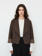 A woman modeling a cropped brown textured jacket with wide lapels and turned-back cuffs, worn over a white top and black trousers against a plain light gray background.