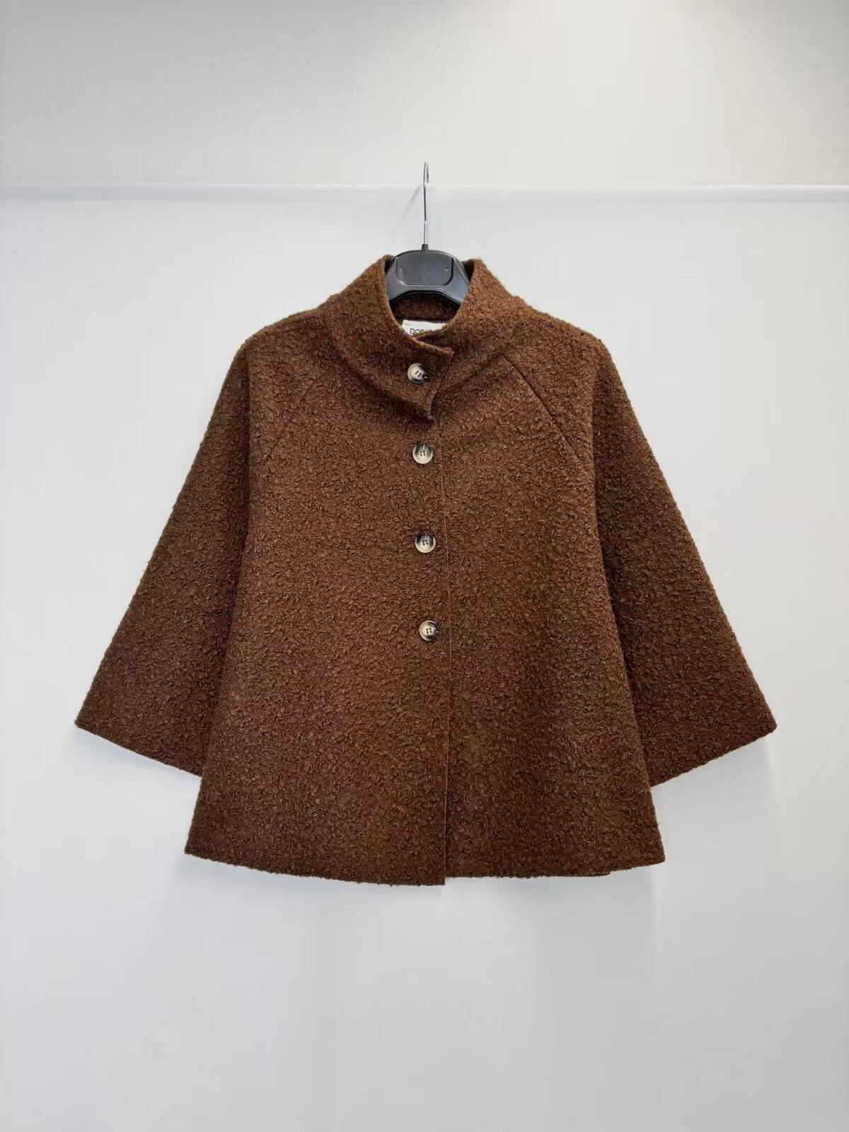 A brown textured bouclé jacket with a mock neck and three front buttons, hanging on a black hanger against a plain white wall.
