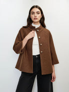 A woman modeling a brown textured bouclé jacket with a mock neck and decorative buttons, worn over a white turtleneck and black wide-leg jeans, against a plain white studio background.