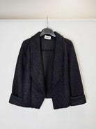 A black textured bouclé jacket with wide lapels and rolled cuffs, hanging on a black hanger against a plain white wall.