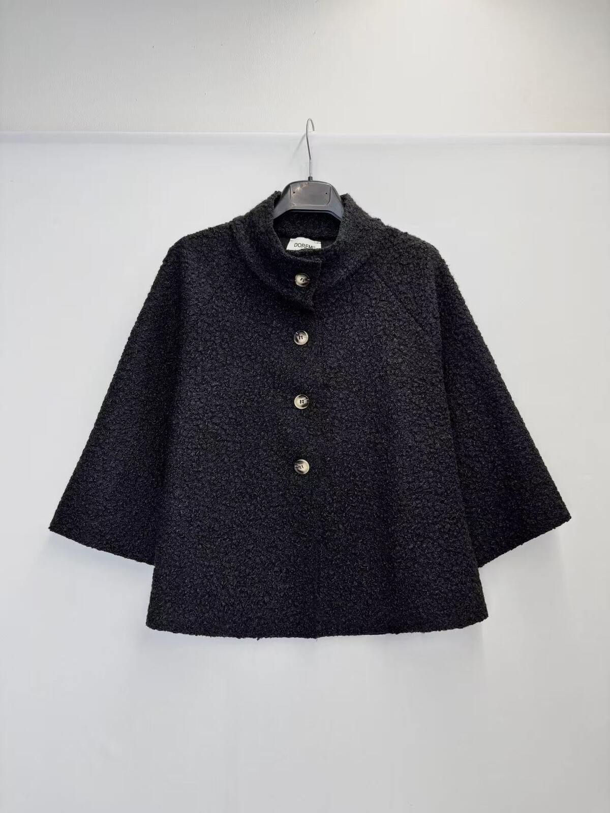 A black textured bouclé cape jacket with a high stand collar and three gold-tone buttons, hanging on a black hanger against a plain white wall.