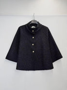 A black textured bouclé cape jacket with a high stand collar and three gold-tone buttons, hanging on a black hanger against a plain white wall.