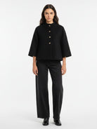 A model stands against a plain white studio background wearing a black cropped cape jacket with three-quarter sleeves and gold-tone buttons, paired with black wide-leg trousers and black shoes.