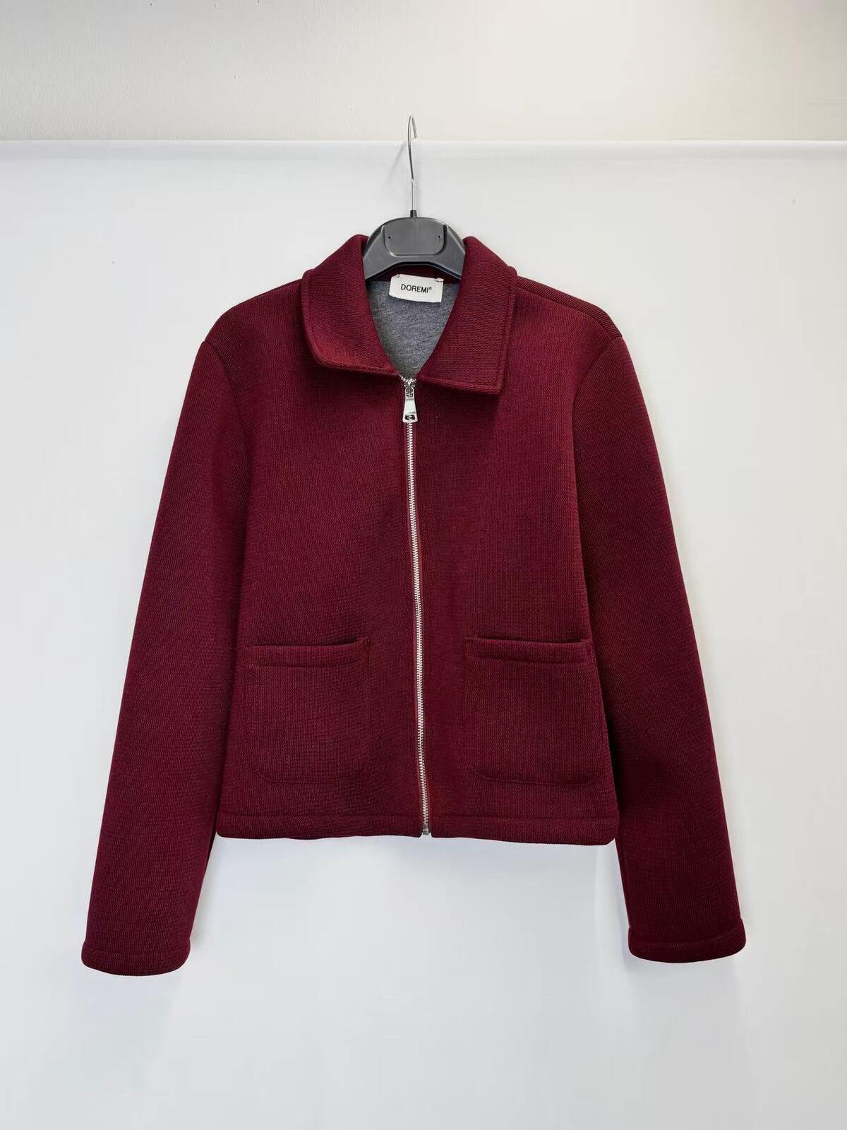 A maroon zip-up jacket with a stand collar and two front patch pockets, hanging on a black hanger against a plain white wall.