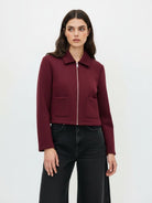 A woman modeling a cropped, long-sleeve zip-up jacket in deep burgundy with two front patch pockets, paired with black wide-leg jeans, against a plain white studio background.