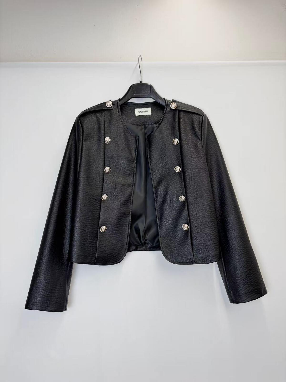 A black cropped faux-leather jacket with silver-tone double-breasted buttons and shoulder epaulets, hanging on a black hanger against a plain white wall.