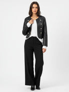 A woman modeling a black leather-look cropped jacket with silver-tone buttons, worn over a white top and black wide-leg trousers, against a plain light gray studio background.