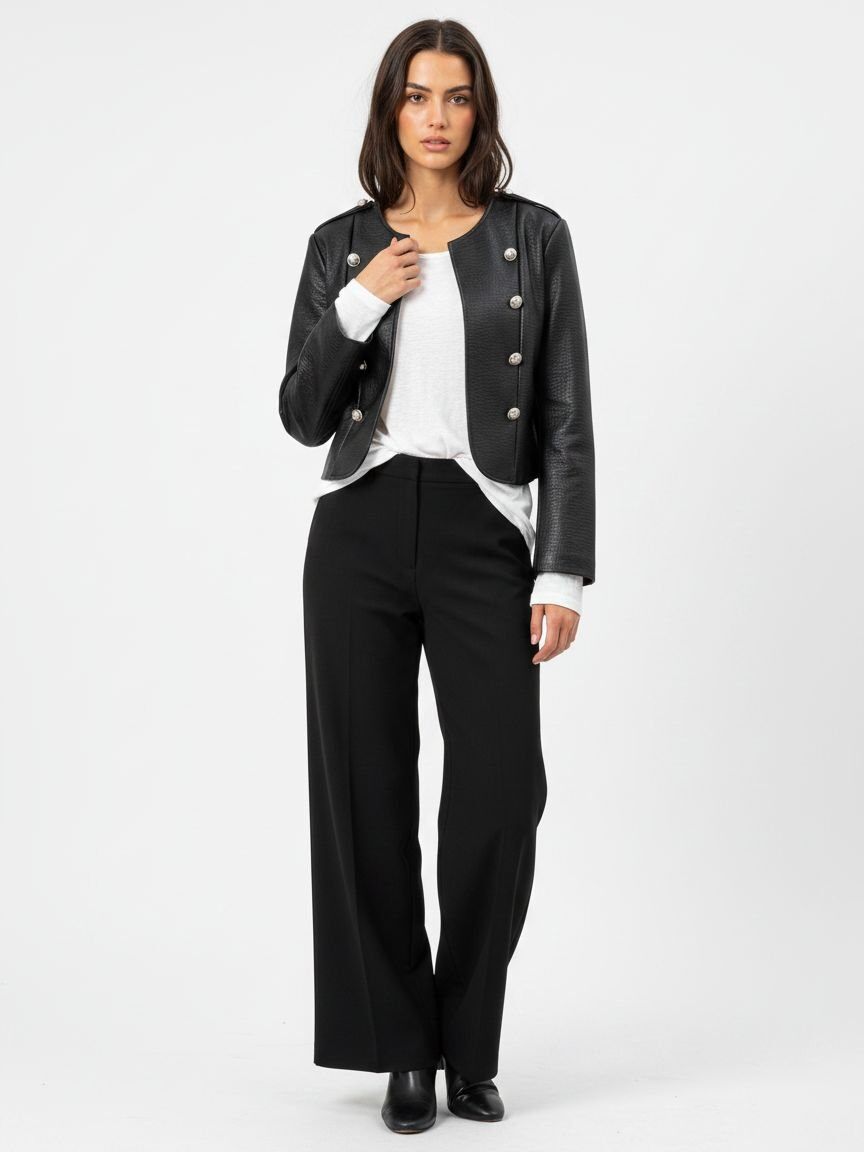 A woman modeling a black leather-look cropped jacket with silver-tone buttons, worn over a white top and black wide-leg trousers, against a plain light gray studio background.