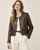 A woman modeling a cropped dark brown faux-leather jacket over a cream top and cream trousers, photographed against a plain light beige studio background.