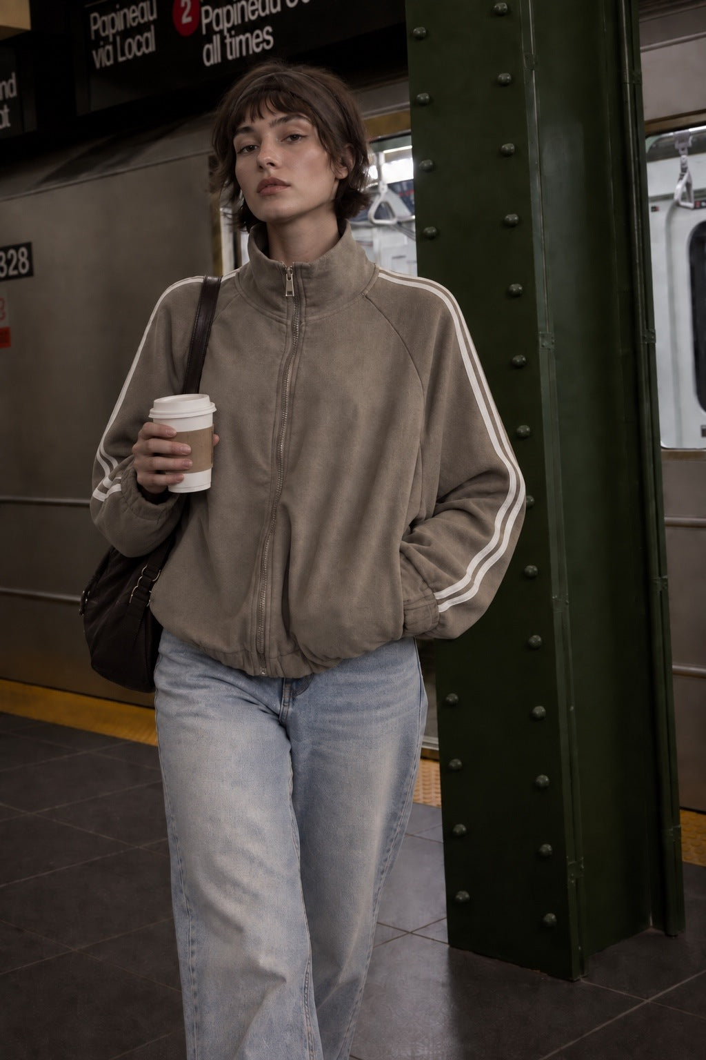 A woman stands in a subway station wearing a taupe-brown zip-up jacket with white stripes on the sleeves, paired with light-wash blue jeans and carrying a black shoulder bag while holding a coffee cup.