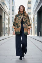 A woman walking down a city street wearing a dark green and brown floral patterned jacket over a black top and wide-leg navy trousers.