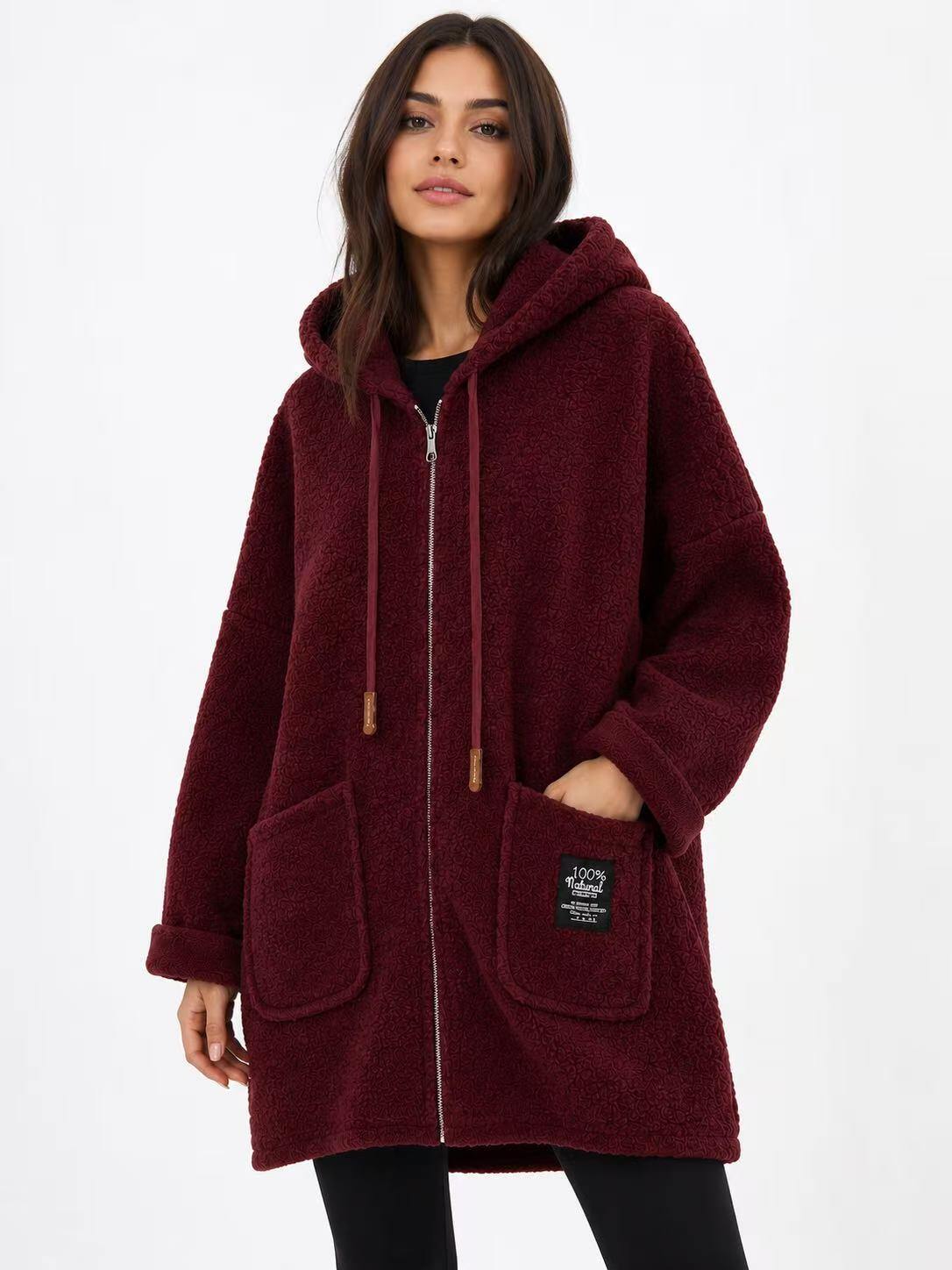 A woman wearing a plush, sherpa-textured hooded zip-up coat in deep burgundy. The coat features a drawstring hood, front pockets, and a small black brand patch on the lower right pocket.