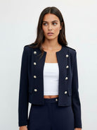 A woman modeling a cropped navy blue double-breasted blazer with silver-tone buttons, worn over a white crop top and matching navy trousers, against a light gray studio background.