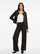 A woman modeling a dark brown cropped double-breasted jacket with buttons, matching dark brown wide-leg trousers, a white tank top, and dark brown boots, set against a plain white studio background.