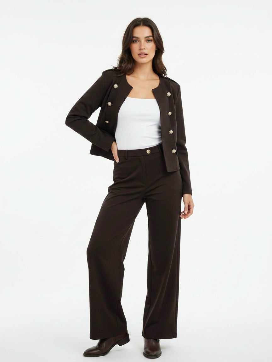 A woman modeling a dark brown cropped double-breasted jacket with buttons, matching dark brown wide-leg trousers, a white tank top, and dark brown boots, set against a plain white studio background.