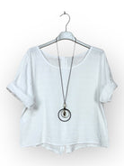 A white short-sleeve linen top hanging on a white hanger, styled with a long black pendant necklace featuring a silver-toned ring and a small gold-toned bead.
