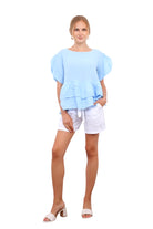 Woman wearing a light blue peplum top and white shorts standing against a white wall