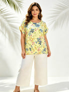 A woman modeling a short-sleeve yellow top with a blue, green, and pink floral print, paired with white cropped pants, set against a white background with palm leaf shadows.