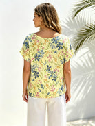 A woman shown from the back wearing a pale yellow short-sleeve top with a blue, green, and pink floral print, paired with white pants